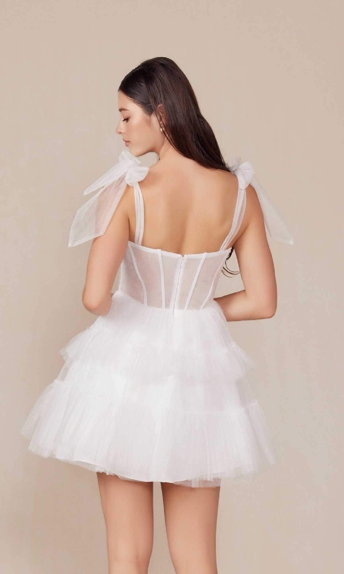 A woman with long dark hair is shown from the back wearing the Sheer-Bodice Short White A-Line Party Dress Y858W, featuring tiered tulle and sheer ribbon straps, against a plain beige background.